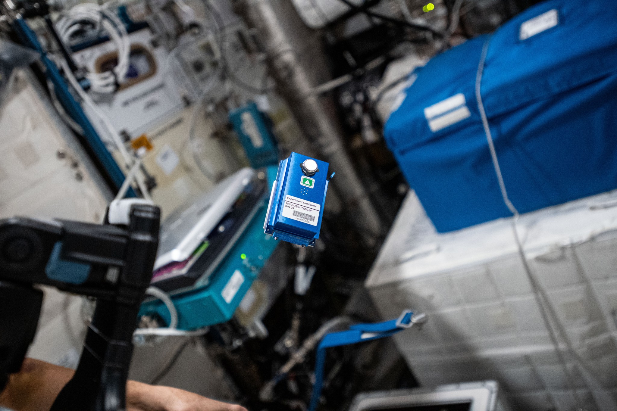 A cell phone-sized blue box with a barcode label and a white button floats in the space station. A closed laptop and several storage boxes are visible in the background and a black camera is mounted in the foreground.