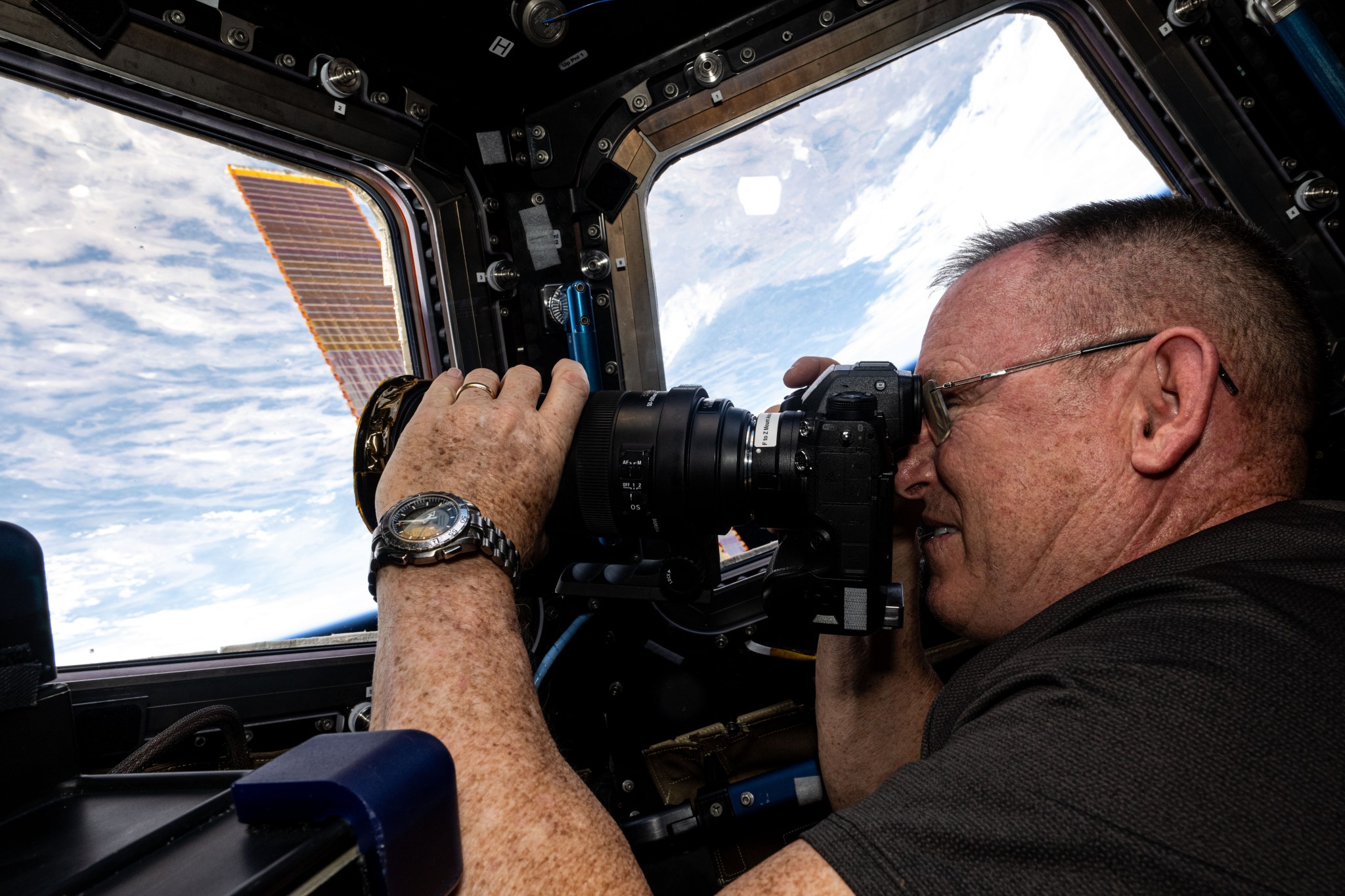 Wilmore, wearing a black polo shirt and a watch on his left wrist, holds a camera to his face with his right hand and adjusts the lens with his left. He is facing one of the cupola windows and a second window is visible to his right. A cloudy Earth below and a bit of one of the station’s solar panels can be seen through the windows.