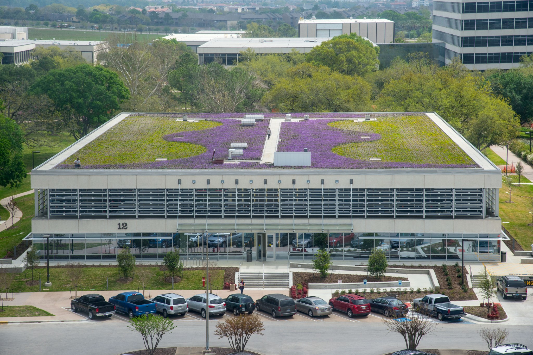 Aerial view of a building with a large rooftop garden featuring a mix of green and purple vegetation. The garden is designed in a curving, artistic pattern. In the background, other buildings and lush greenery are visible.