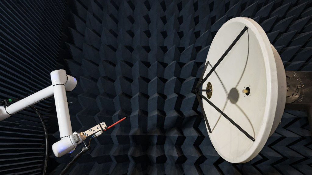 In a testing facility, a robotic arm manipulates a probe to test the electric field of a large, white, circular dish antenna. This antenna is situated within an anechoic chamber, which is lined with dark blue, wedge-shaped foam.