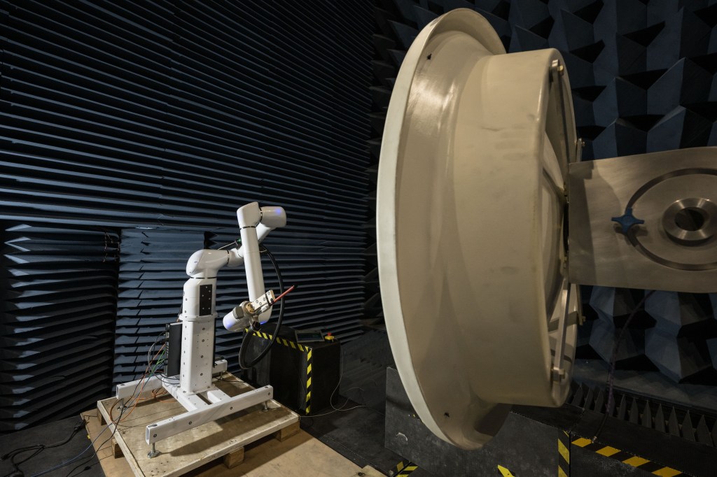 In a testing facility, a robotic arm manipulates a probe to test the electric field of a large, white, circular dish antenna. This antenna is situated within an anechoic chamber, which is lined with dark blue, wedge-shaped foam.