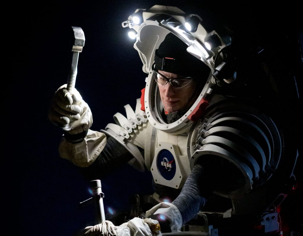 NASA astronaut wearing a mockup spacesuit system, illuminated by the lights on her helmet as she hammers a drive tube into the ground. Civil Space Challenges