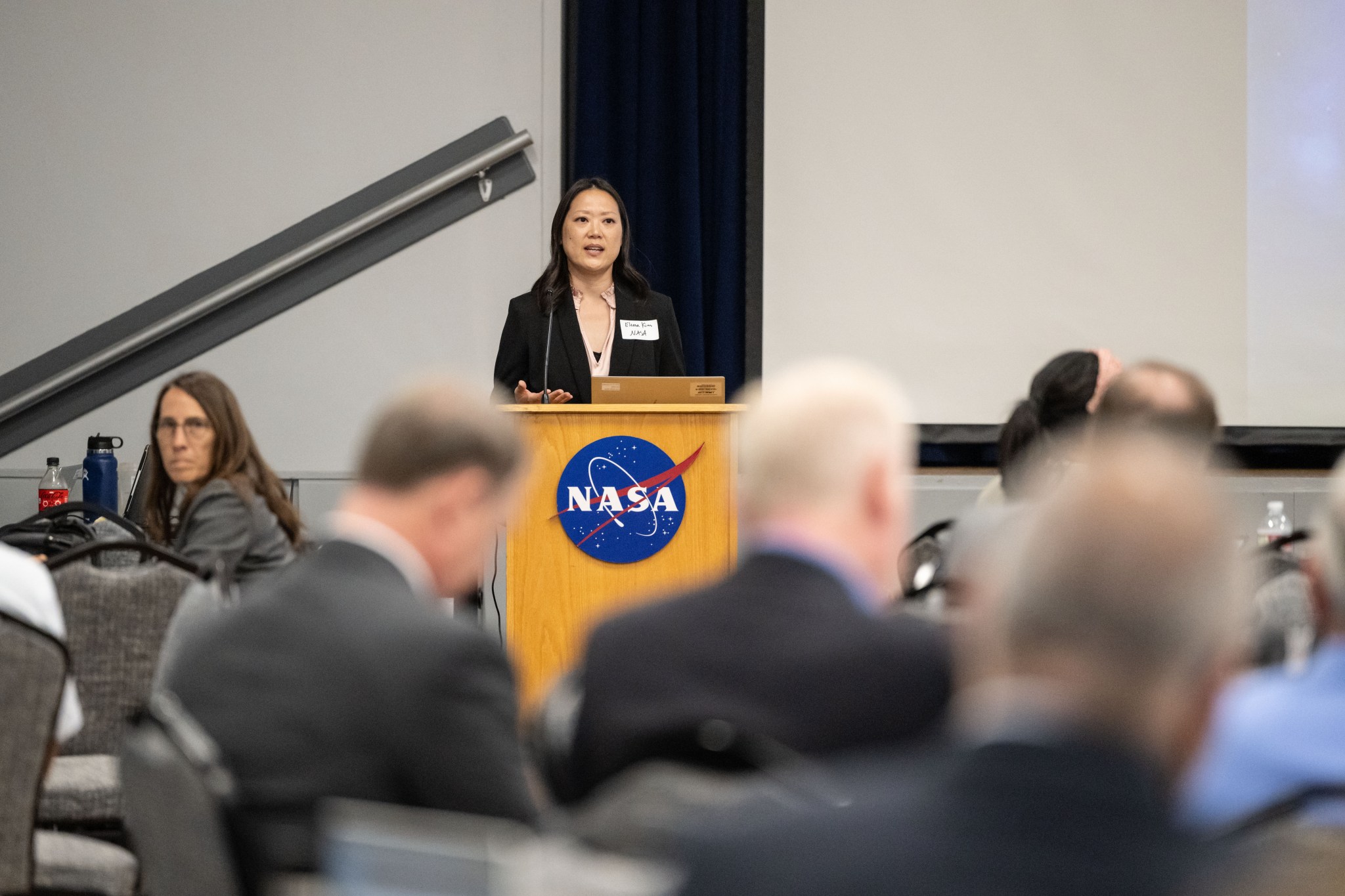 A woman wearing a pink blouse and a black blazer stands at a podium with the NASA meatball logo attached to the front. The woman is speaking to a group of people.