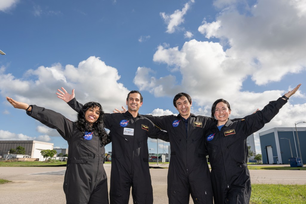 Four NASA personnel in black jumpsuits stand outside and smile with their arms outstretched. The background features a bright blue sky with scattered clouds and some buildings.