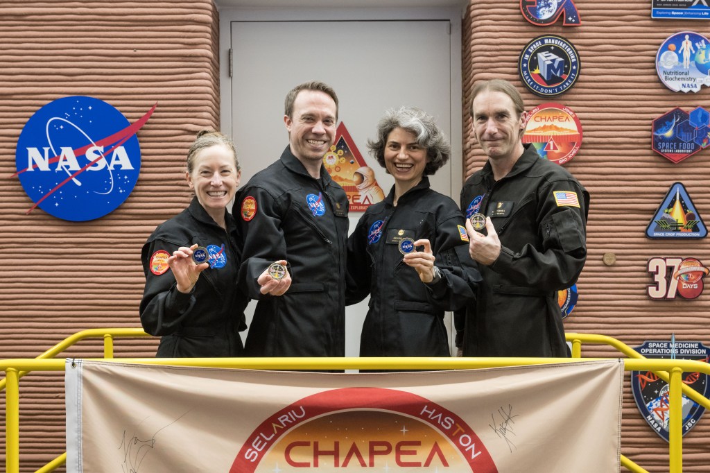 Four people in black NASA jumpsuits are standing in front of a building with NASA logos and mission patches on the wall. They are smiling and holding mission patches, posing for a group photo.