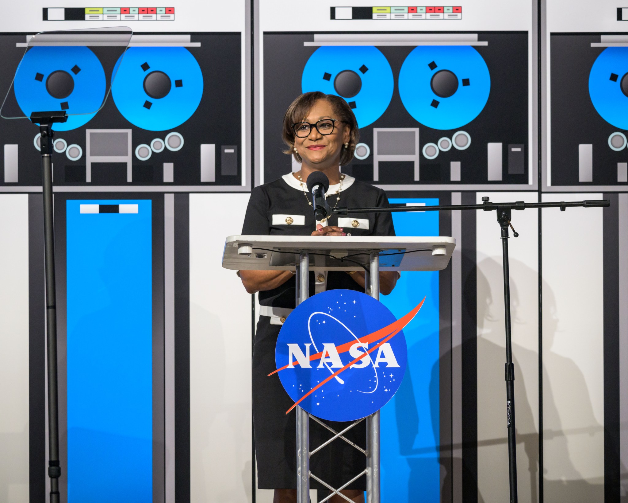 A woman stands at a podium with the NASA logo, delivering a speech. She is dressed in a black and white outfit and wearing glasses. Behind her is a backdrop featuring large images of tape reels and computer equipment. Two microphones and a teleprompter are set up on either side of the podium.