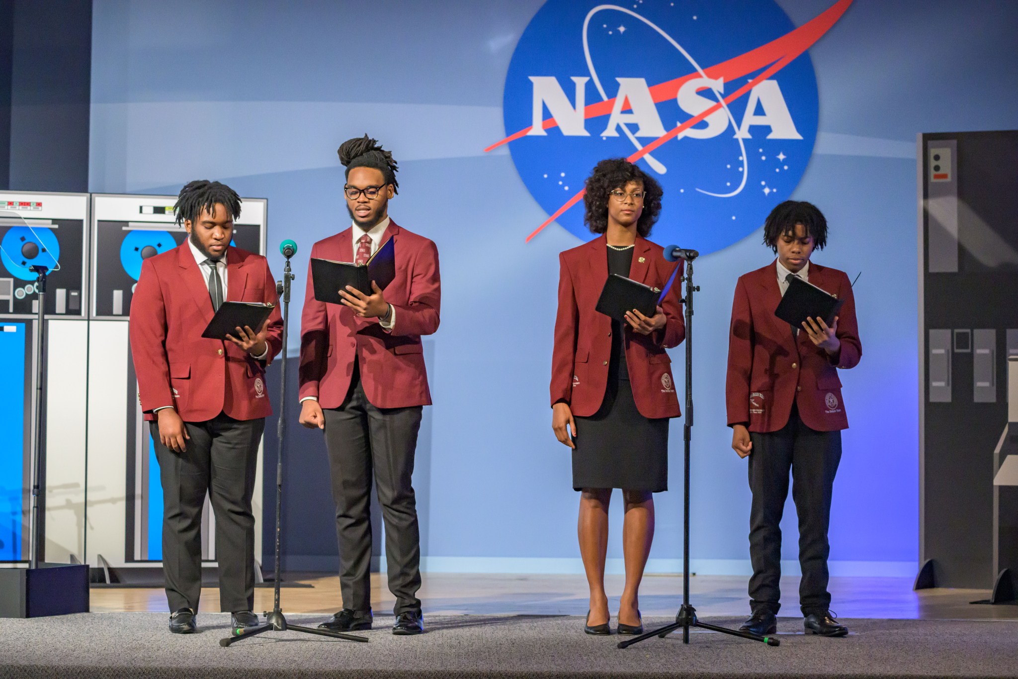 Four young people stand on a stage in front of a large NASA logo. They are dressed in matching maroon blazers and are holding black folders.The backdrop features large images of tape reels and computer equipment.