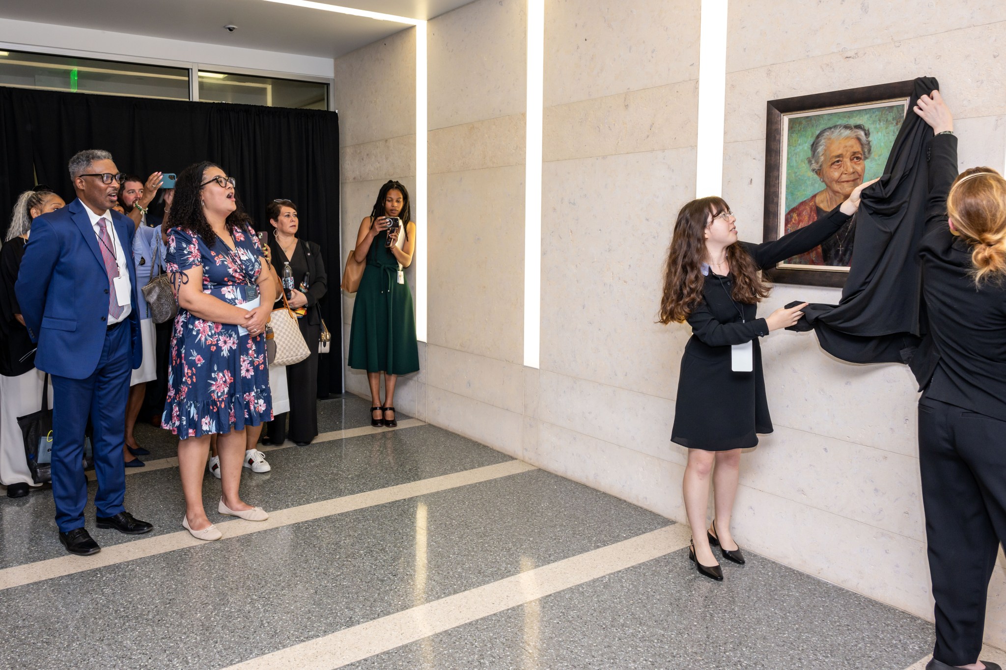 A group of people gather indoors for the unveiling of a portrait. Two women, one young and one older, are removing a black cloth to reveal a framed painting of an elderly woman. The attendees, including a man in a blue suit and a woman in a floral dress, watch the unveiling.