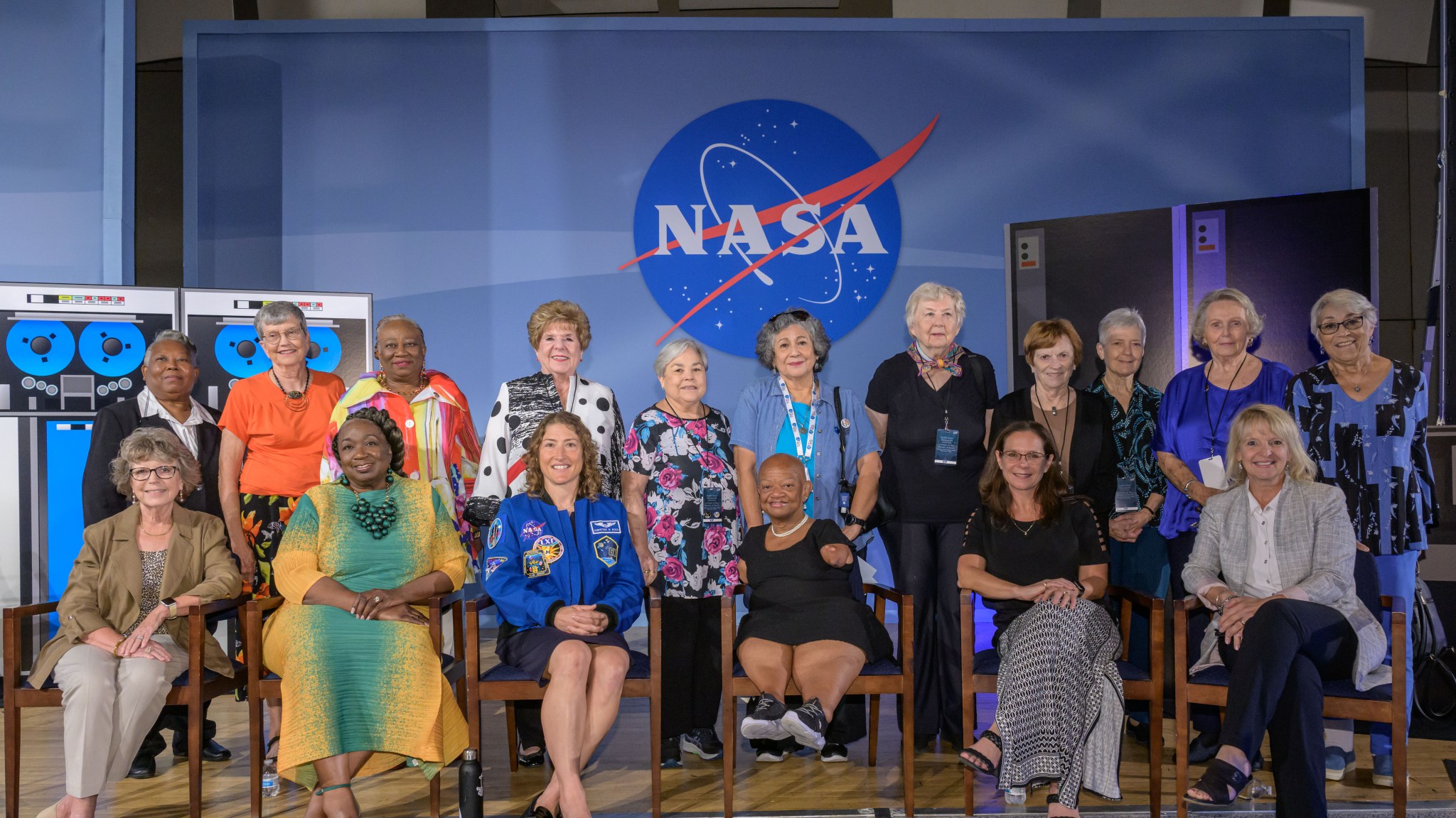A group of women pose for a photo in front of a large NASA logo backdrop. The group includes women of various ages, some seated and some standing. They are dressed in a variety of outfits, from professional attire to casual wear.