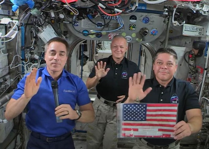 July 4, 2020. Expedition 63 astronauts Christopher J. Cassidy, left, Douglas G. Hurley, and Robert L. Behnken, hold the Legacy Flag flown on STS-1 and STS-135