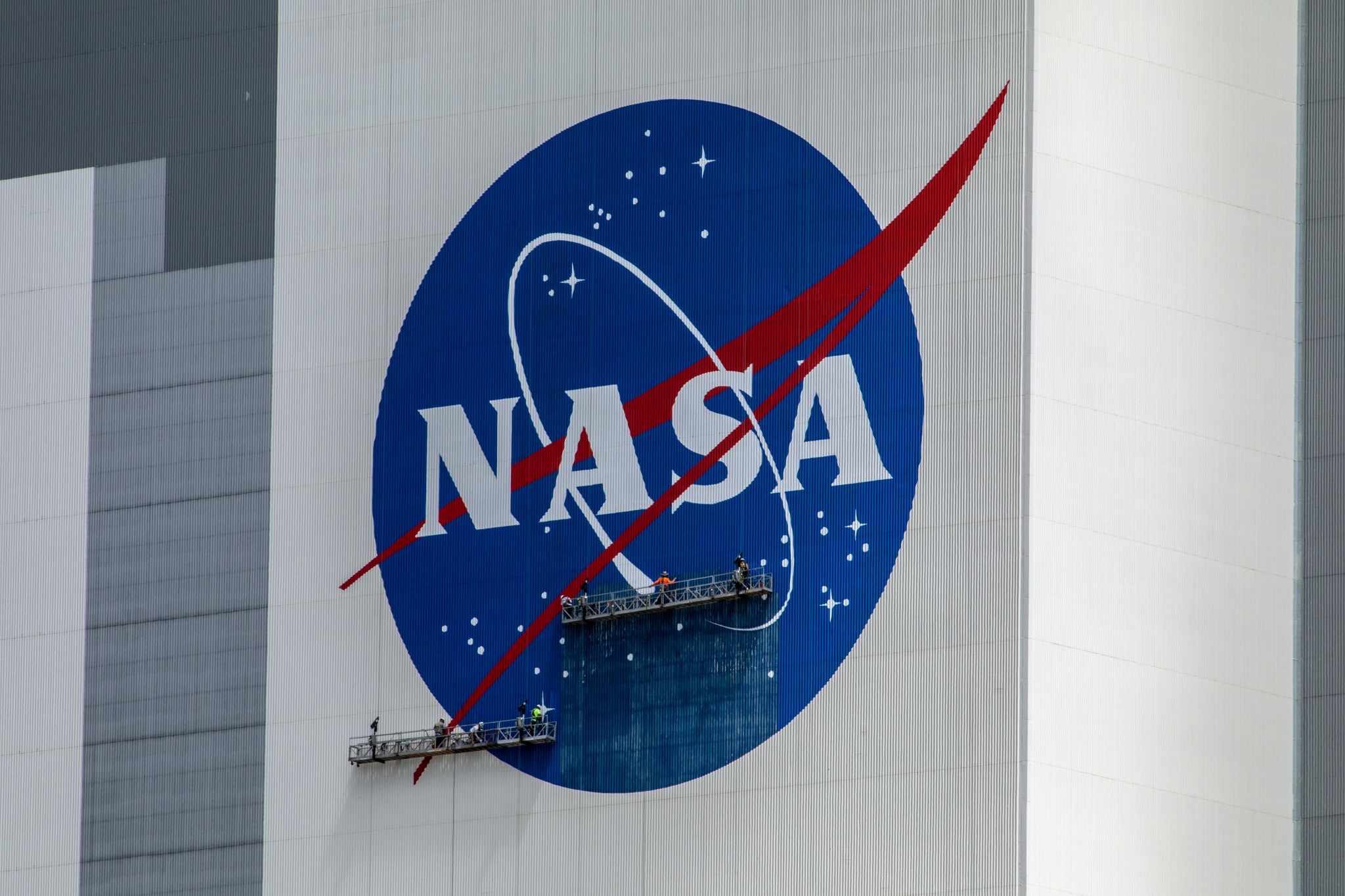 Painters stand on suspended platforms as they work on painting the red, white, and blue NASA "meatball" logo on the white and gray Vehicle Assembly Building. The painters are tiny compared to the size of the logo.