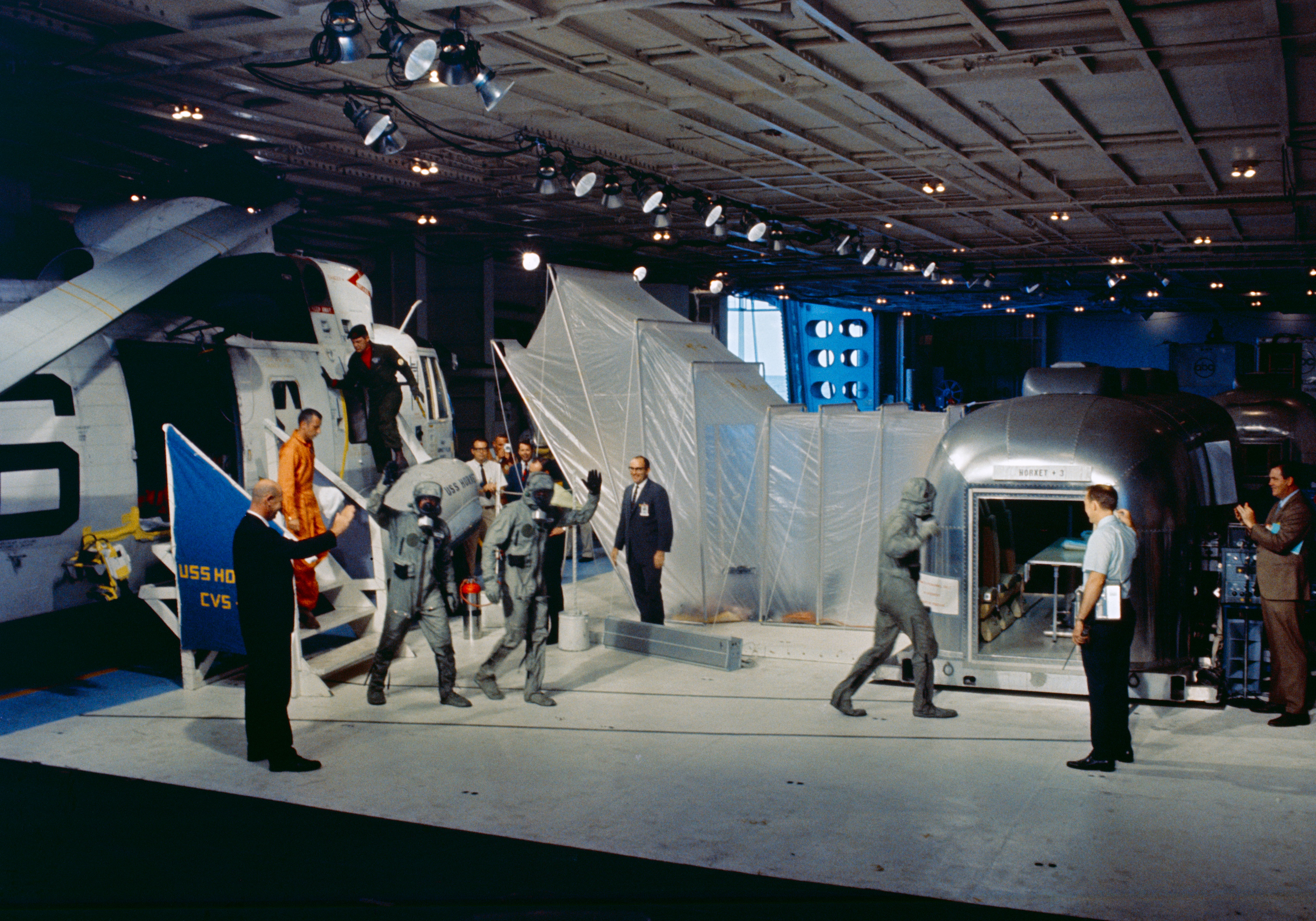 Once aboard the U.S.S. Hornet, Mike, Neil, and Buzz wearing their BIGs walk the 10 steps from the Recovery One helicopter to the Mobile Quarantine Facility (MQF), with NASA flight surgeon Dr. William Carpentier, in orange suit, following behind