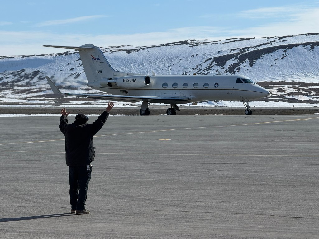 NASA's white Gulfstream III aircraft is seen on the runway taxiing. A flight crew member has his arms raised helping direct the pilots as they prepare to take off. The bottom of the photo is flat, gray concrete. Low, snow covered hills can be seen behind the aircraft.