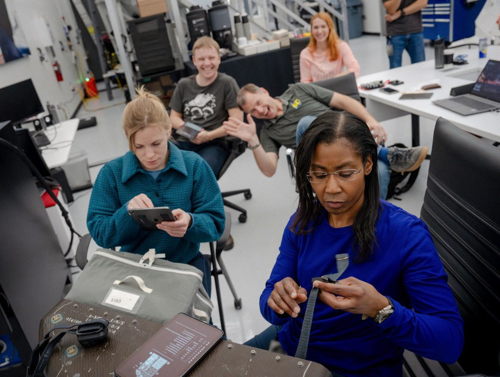 NASA’s SpaceX Crew-9 members Mission Specialist Aleksandr Gorbunov and Pilot Nick Hague smile big for the camera while Commander Zena Cardman and Mission Specialist Stephanie Wilson train on how to route straps and buckles around various bags that need to be stored in the Dragon spacecraft.