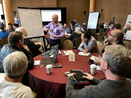 A round table of people looking to the person who is standing up and talking