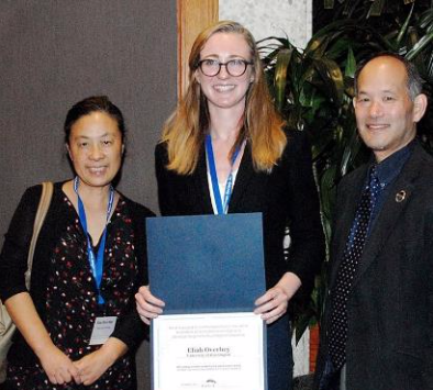 Two females and one male posed together smiling. The female in the middle is holding an award certificate.