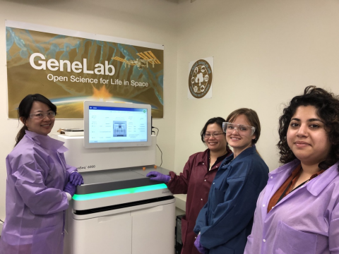 4 women smiling and standing by the sample processing machine with lab coats on and some with protective eyewear.