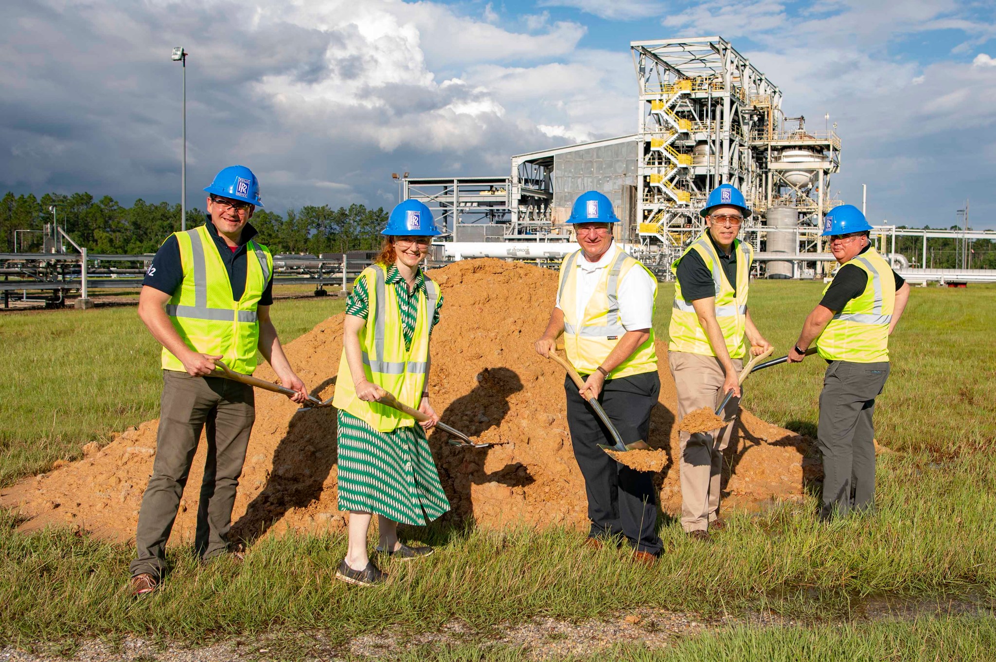 five officials wearing blue hard hats and yellow vests dig into red clay dirt at the site of a new Rolls-Royce test pad at Stennis Space Center