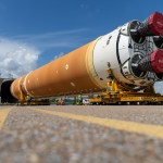 The core stage of the Artemis Space Launch System being loaded on a covered barge. The stage is a large cylinder shape with the engines facing toward the camera on two yellow transporters that are guiding the stage into a covered grey container in the background. The body of the cylinder is mostly an orange color and white around the bottom. The four engines on the bottom are covered with red material.