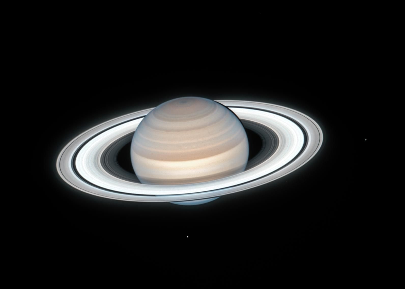 Saturn and its rings appear to glow against the darkness of space. At right and at middle bottom, two small dots can be seen: they are two of Saturn's moons - Mimas and Enceladus, respectively. Saturn itself takes on a reddish tint in its northern hemisphere and blue at its bottom. The planet's concentric rings are an icy white.