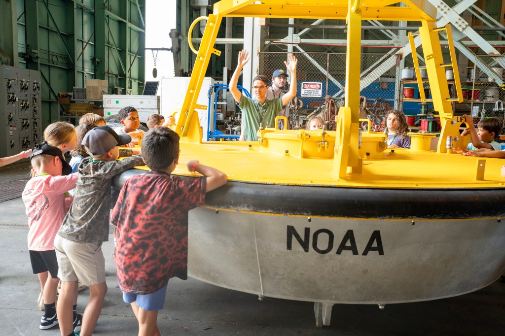 Take Our Children to Work Day participants surround a yellow NOAA buoy as Brandon Elsensohn conducts an activity