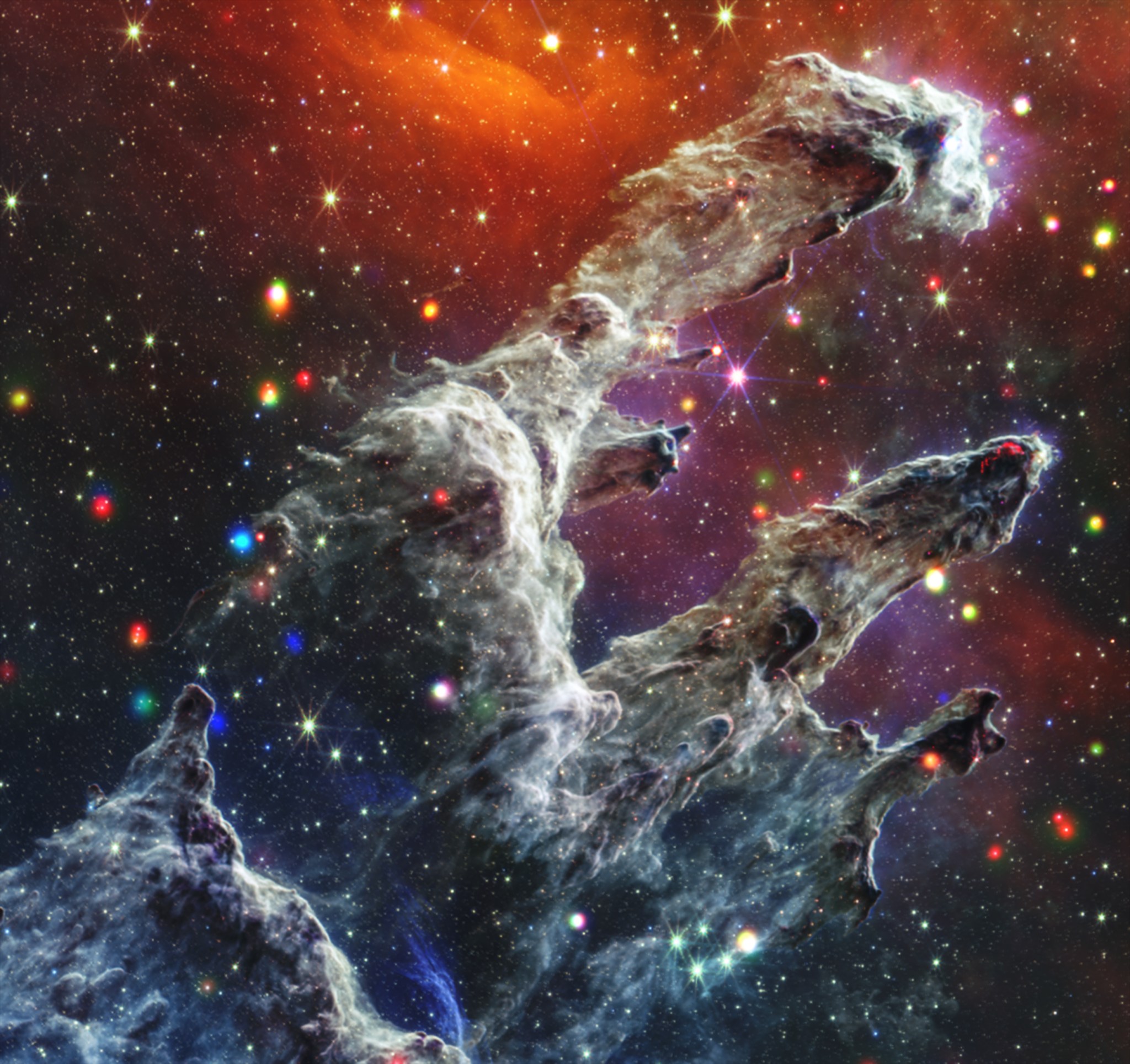 This composite image features a region of star formation known as the Pillars of Creation. Here, tall columns of grey gas and dust emerge from the bottom edge of the image, stretching toward our upper right. Backed by dark orange and pink mist, the cloudy grey columns are surrounded by dozens of soft, glowing, dots in whites, reds, blues, yellows, and purples. These dots are young stars emitting X-ray and infrared light. Churning with turbulent gas and dust, the columns lean to our right with small offshoots pointing in the same direction. The misty glow, colorful stars, and lifelike grey dust formations combine to create an image of yearning cloud creatures at dusk, reaching for something just out of frame.