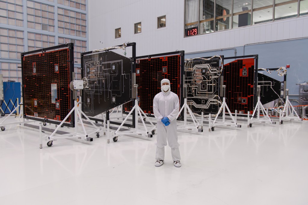 Aaron Vigil stands in front of spacecraft hardware in the Goddard cleanroom. He wears a full cleanroom suit.