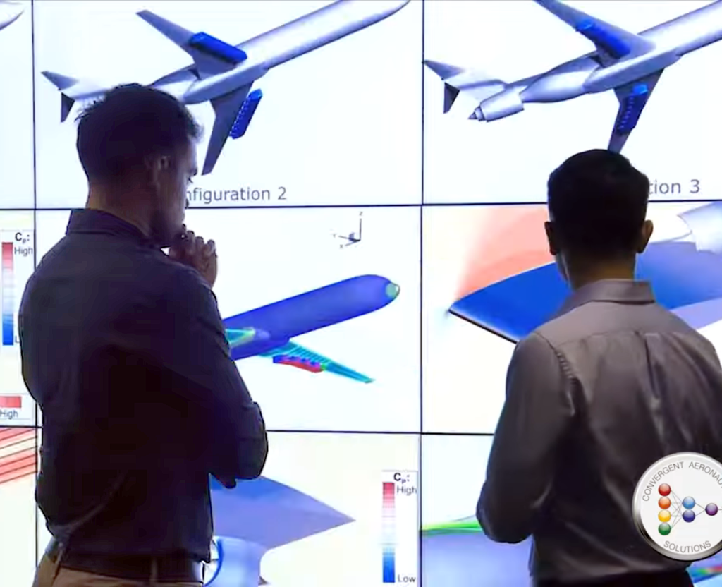 Two men facing away from the camera look at a large video screen showing computer simulations of aircraft.