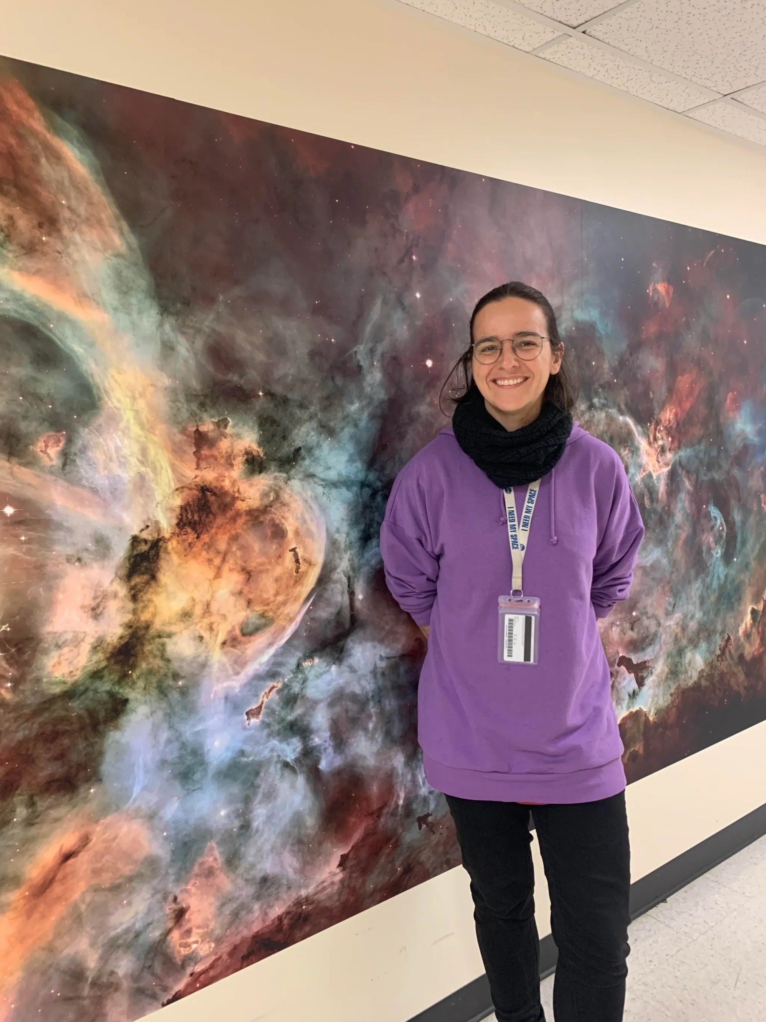 Ariadna Farrés-Basiana stands in front of a colorful Hubble image in a hallway wearing a purple hoodie and black pants. 