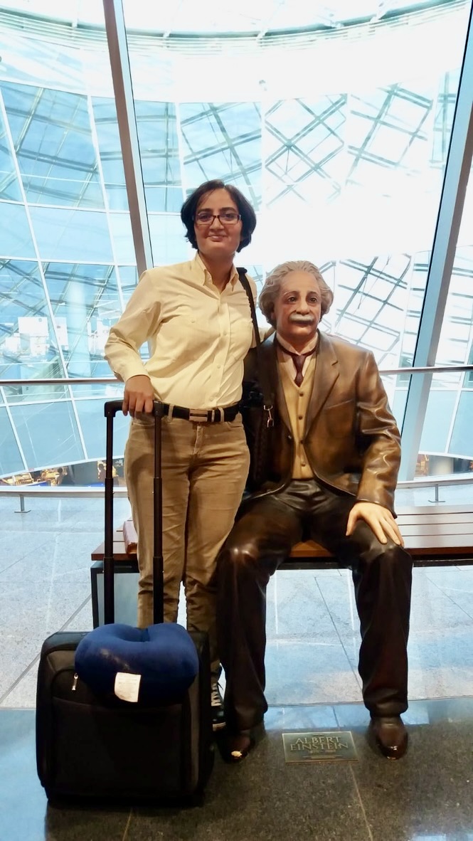 Bindu Rani stands in in front of glass windows next to a statue of Albert Einstein. She is wearing a light colored button down with tan pants and is holding the handle to a blue suitcase. 