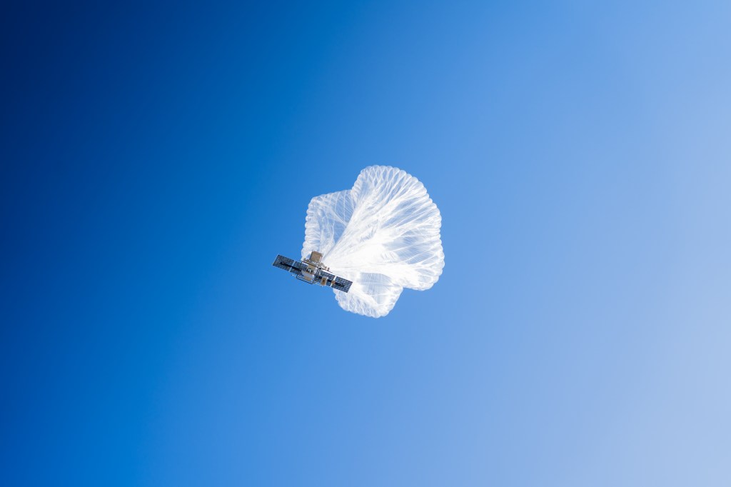 A high-altitude balloon viewed from below against a bright blue sky while carrying a payload.