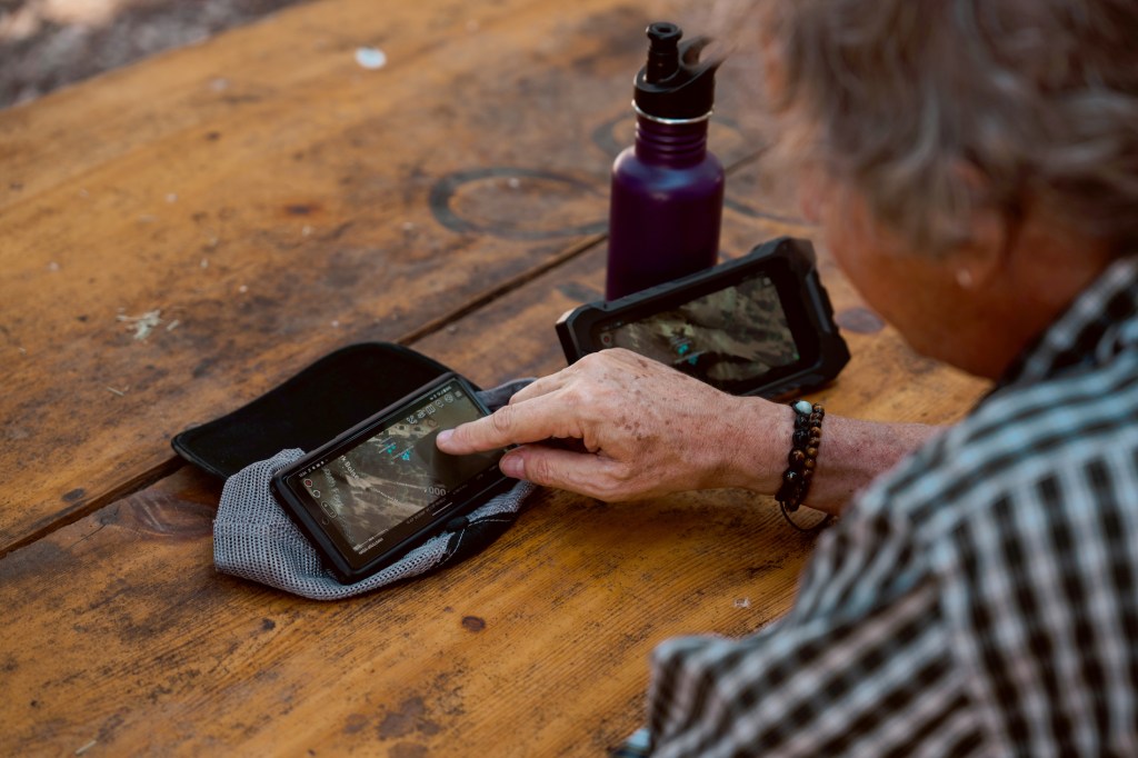 A small handheld mobile device sits on a wooden table. A person is touching the screen.