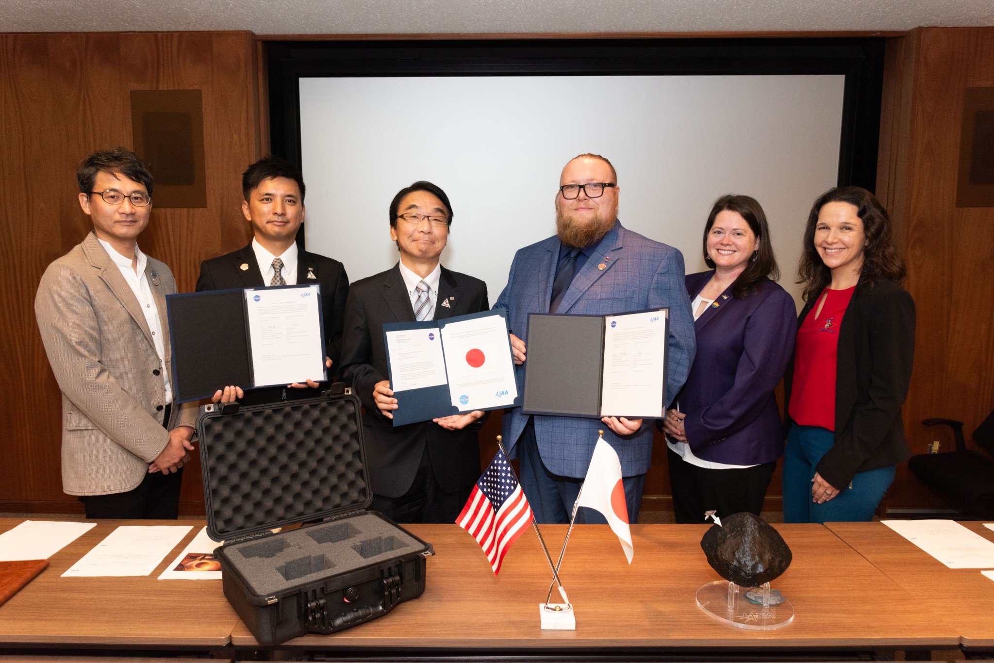 A group of six individuals, consisting of three men and three women, are standing together holding signed documents. The men are on the left and center, wearing business suits, and the women are on the right.