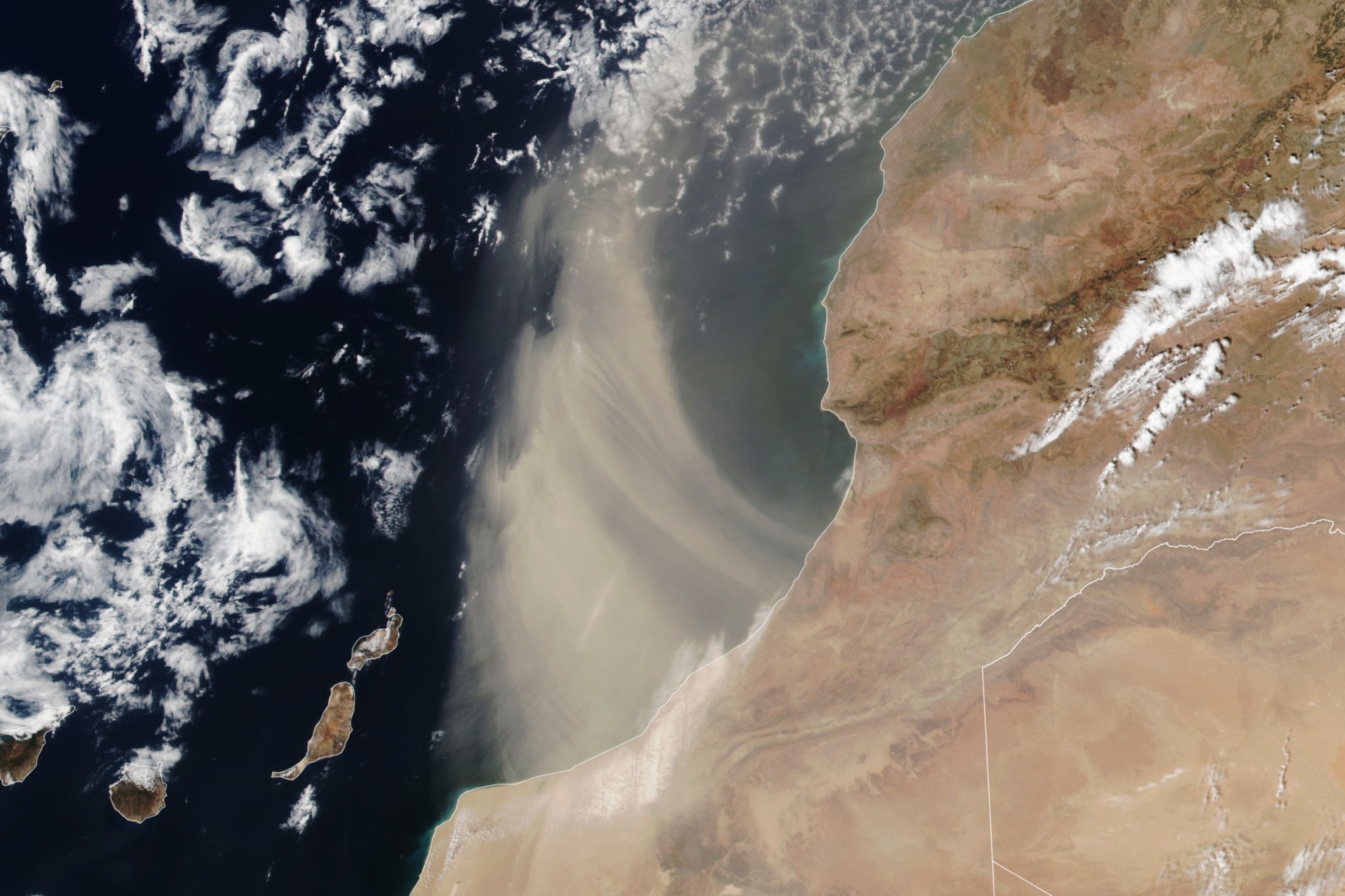 At center, light brown sand wafts over the dark blue ocean. Some white clouds are over the ocean at left and a brown land mass is on the right.