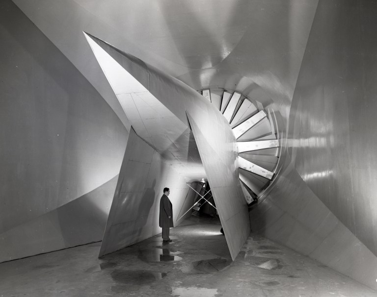 A black-and-white photo of the inside of the Icing Research Tunnel. A man stands beneath the back end of a massive fan.