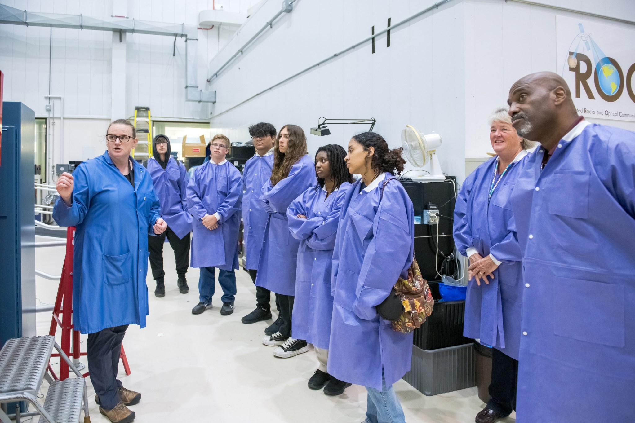 NASA subject matter expert talks with students in lab coats, right, about equipment in the ISS Payload Operations Center.