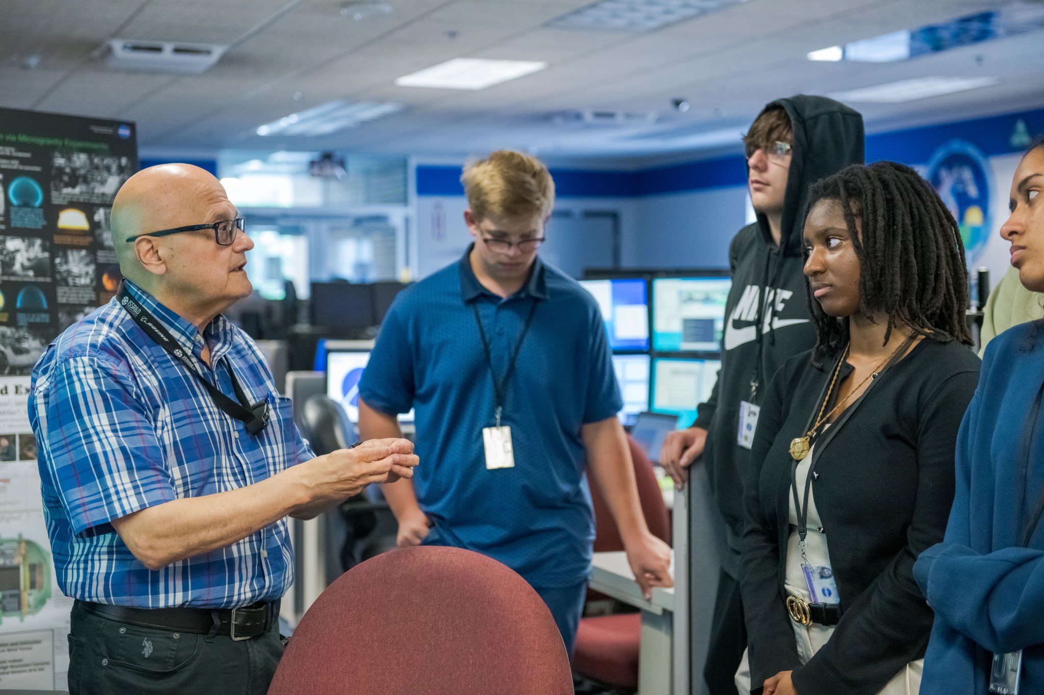 A NASA subject matter expert, left, explains details to four students about the ISS Payload Operations Center. Several monitors tracking experiments are in the background.