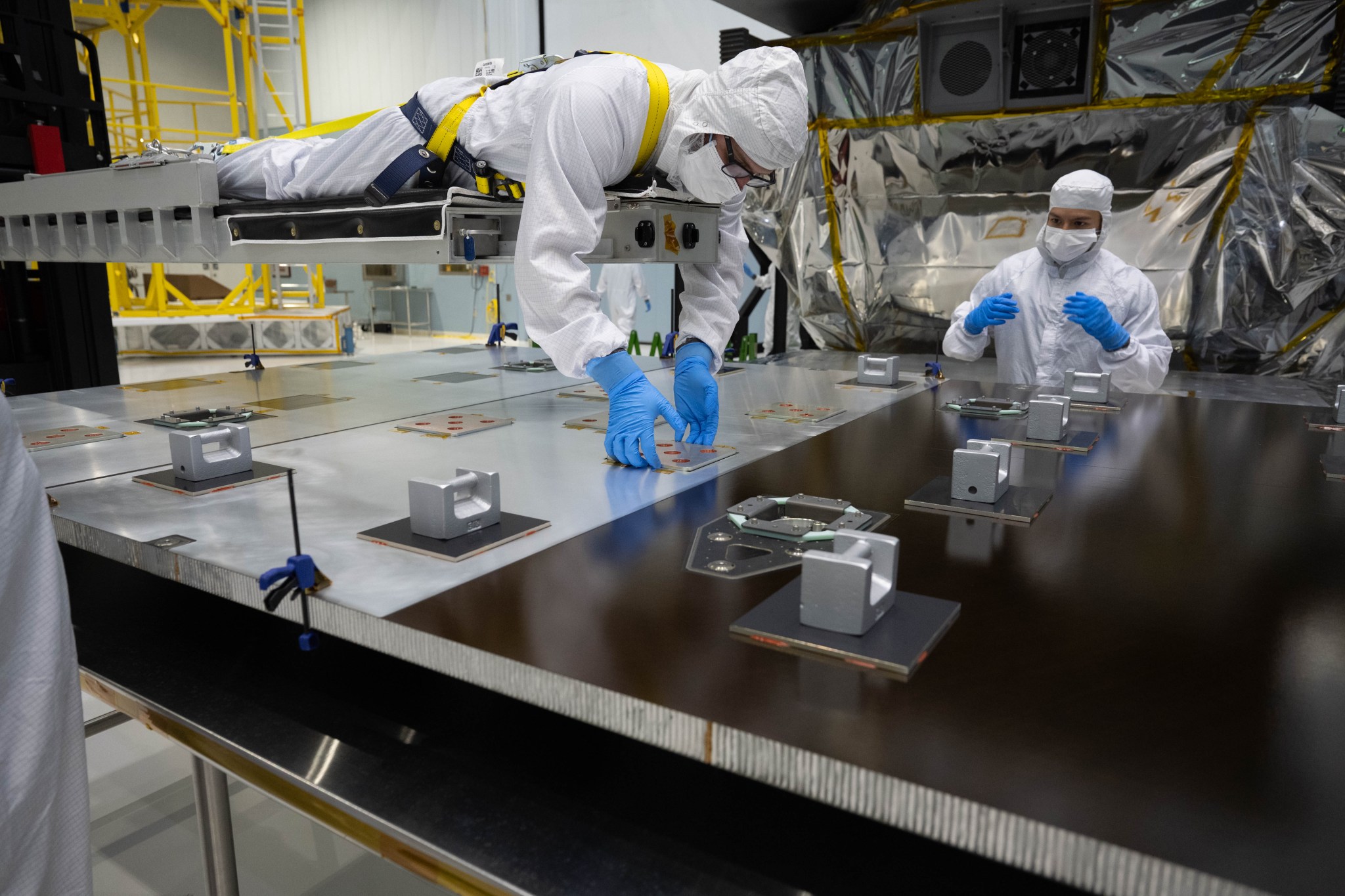 Aaron Vigil lays on a structure above a table to perform to work on a spacecraft part. He is fully dressed in cleanroom gear including a white clean room suit and blue gloves. A yellow harness straps him to the structure he lays on to work above the table.