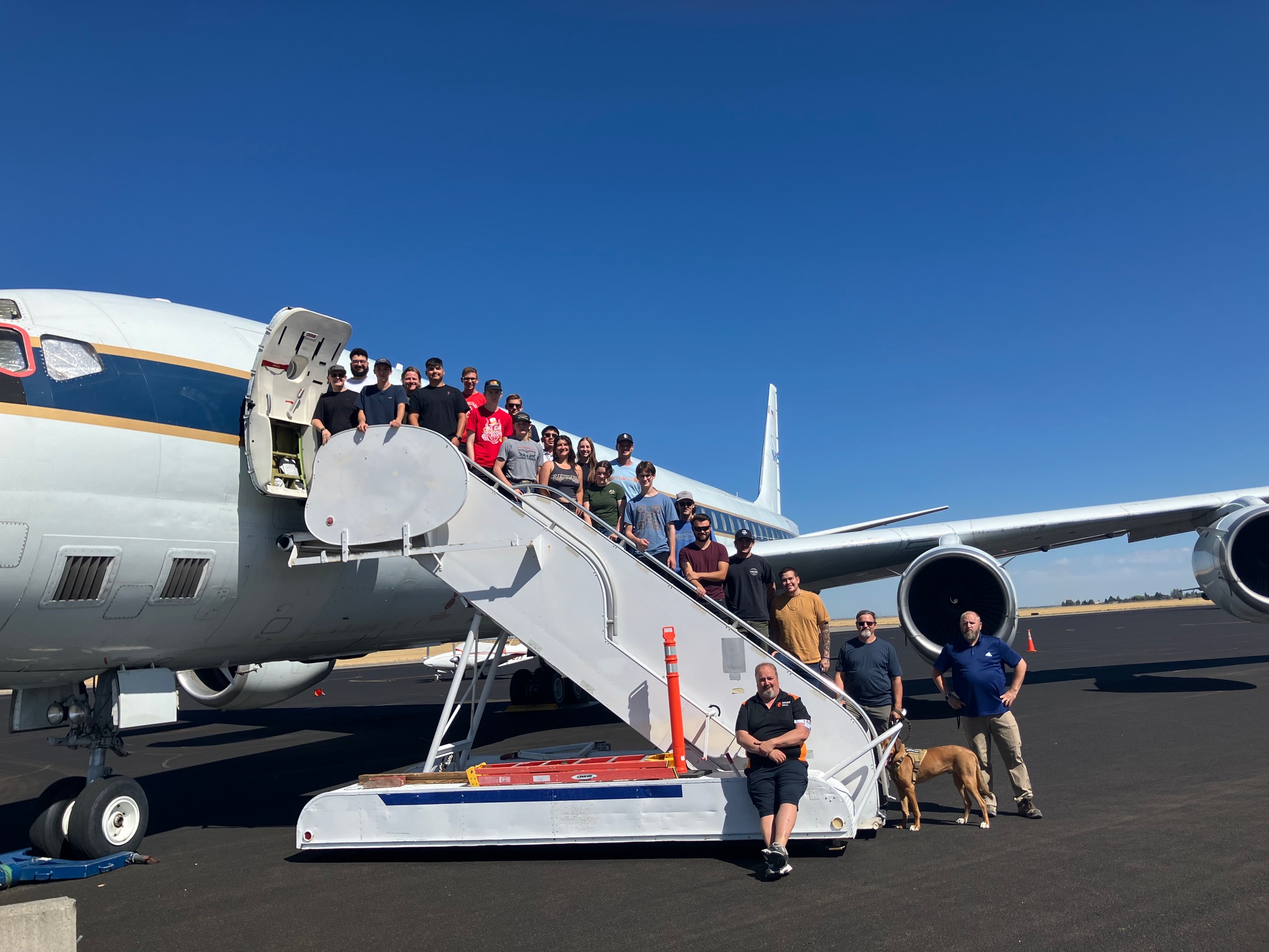 A group of 22 students pose on a stairway descending from an aircraft door. The plane and stairway are white, on a sunny tarmac.