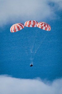 A spacecraft descends towards Earth under three large, red-and-white parachutes. The parachutes are fully deployed.