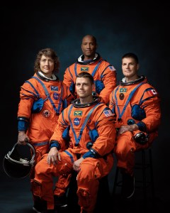 Four people in orange and blue space suits pose in front of a blue backdrop.