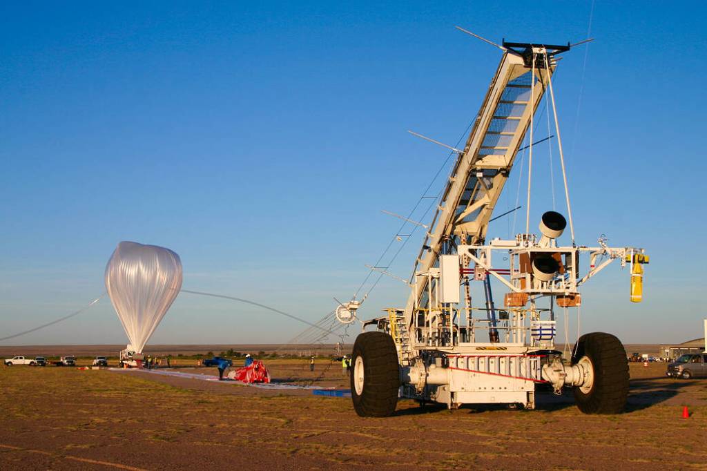 A crane holds a science instrument as a large scientific balloon, tethered to the ground, inflates before liftoff in New Mexico.