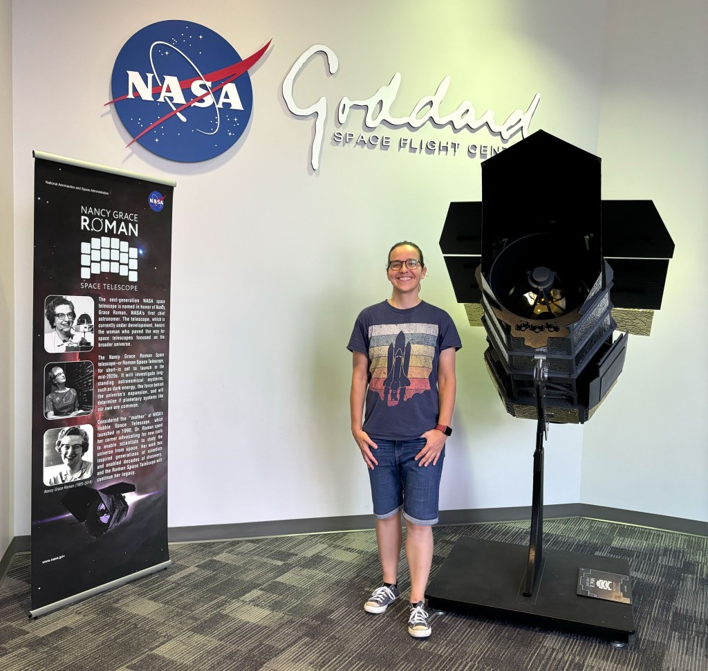 Dr. Ariadna Farrés-Basiana stands in between a model of the Nancy Grace Roman Space Telescope and sign showing history of the telescope. She is wearing a t shirt with a space shuttle graphic and jean shorts. The NASA meatball and Goddard Space Flight Center logo is on the wall behind her.
