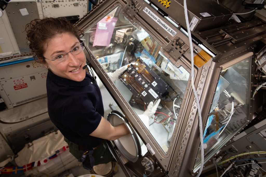 An astronaut wears a headset and glasses while working with scientific equipment aboard the International Space Station. She is manipulating the experiment through glove ports, and various instruments and cables are visible around her in the laboratory module.