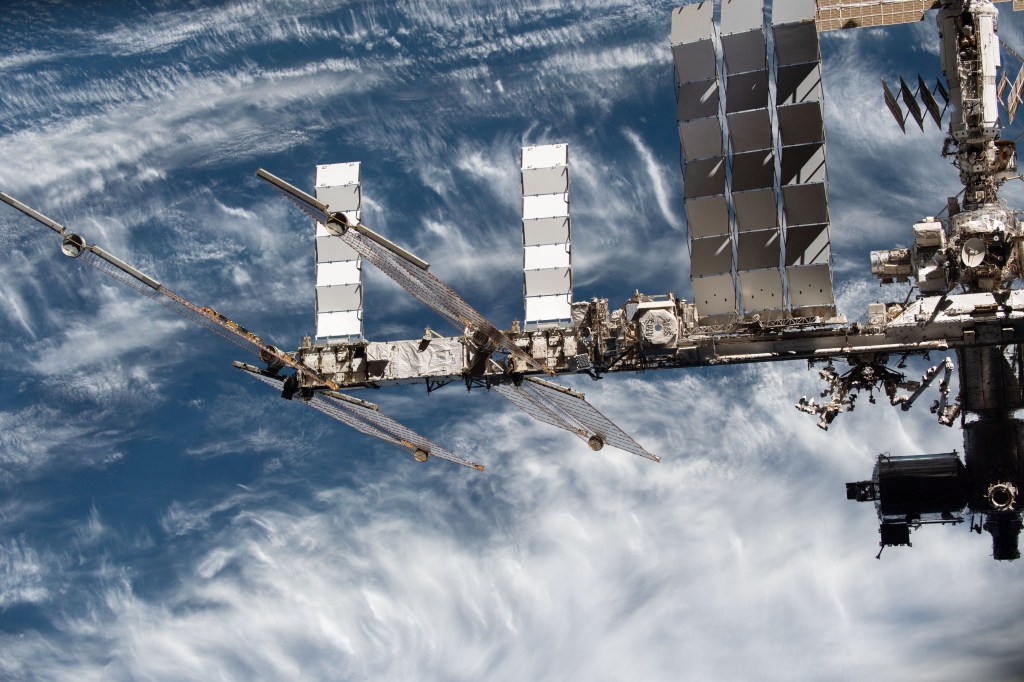 A truss of the space station extends from the right across the center of this image. Several modules of the station are visible on the right. From right to left are a large solar panel extending toward the top of the image, and next to it, the hexagonal top of AMS-02 is visible. Two additional solar panels extend toward the top of the image and two others to either side of the truss. In the background is the blue Earth with scattered white clouds.