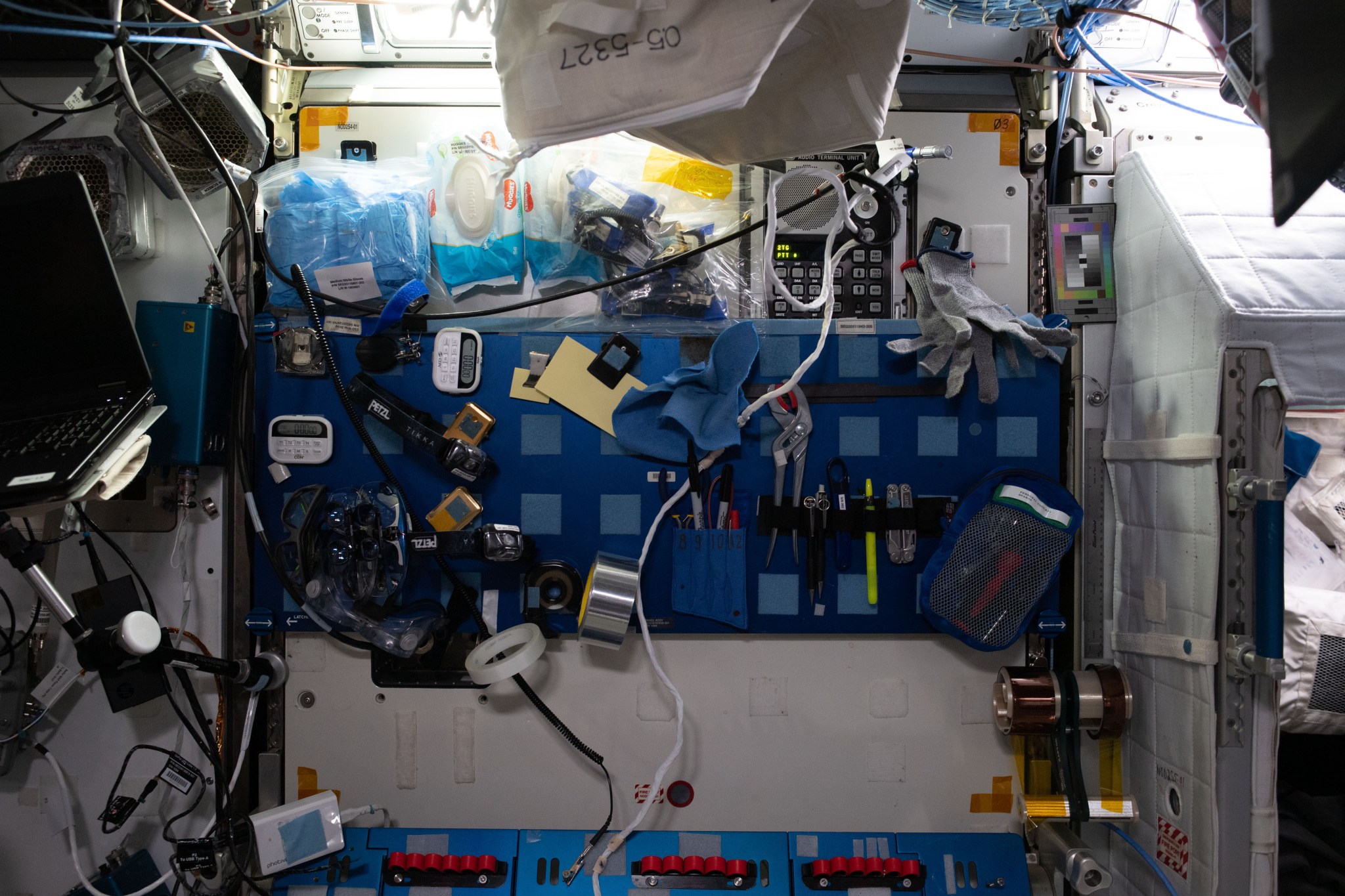 A cluttered workstation in the International Space Station, featuring a variety of tools and equipment secured to a blue panel with Velcro strips.