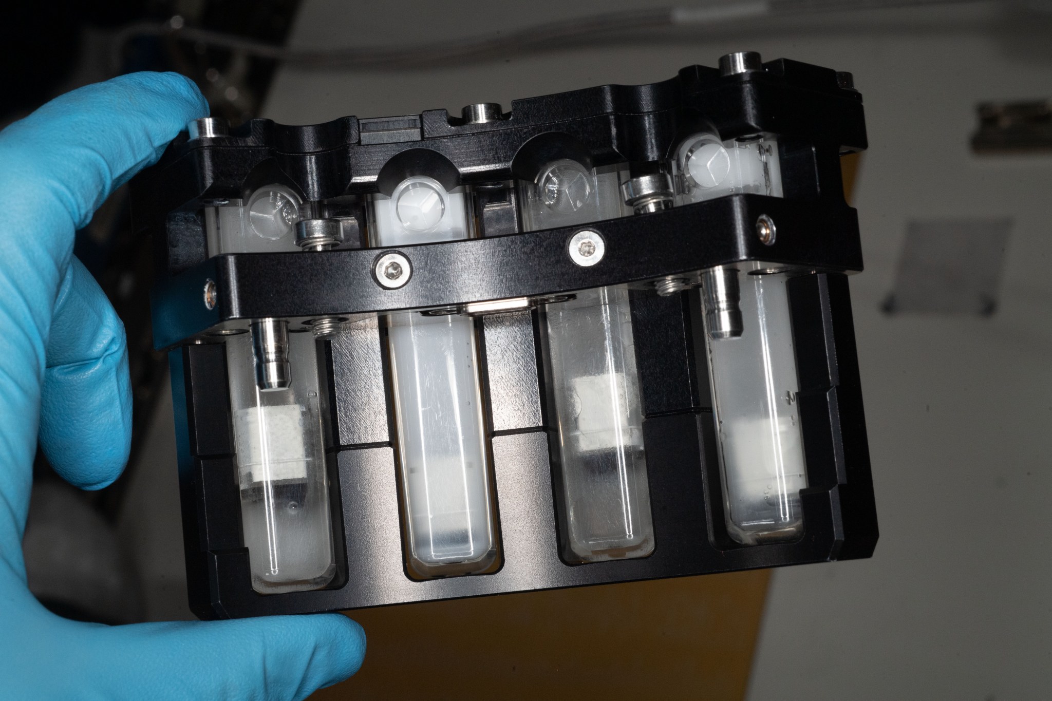 Picture of a latex glove-covered hand holding a black container with four clear tubes inset.