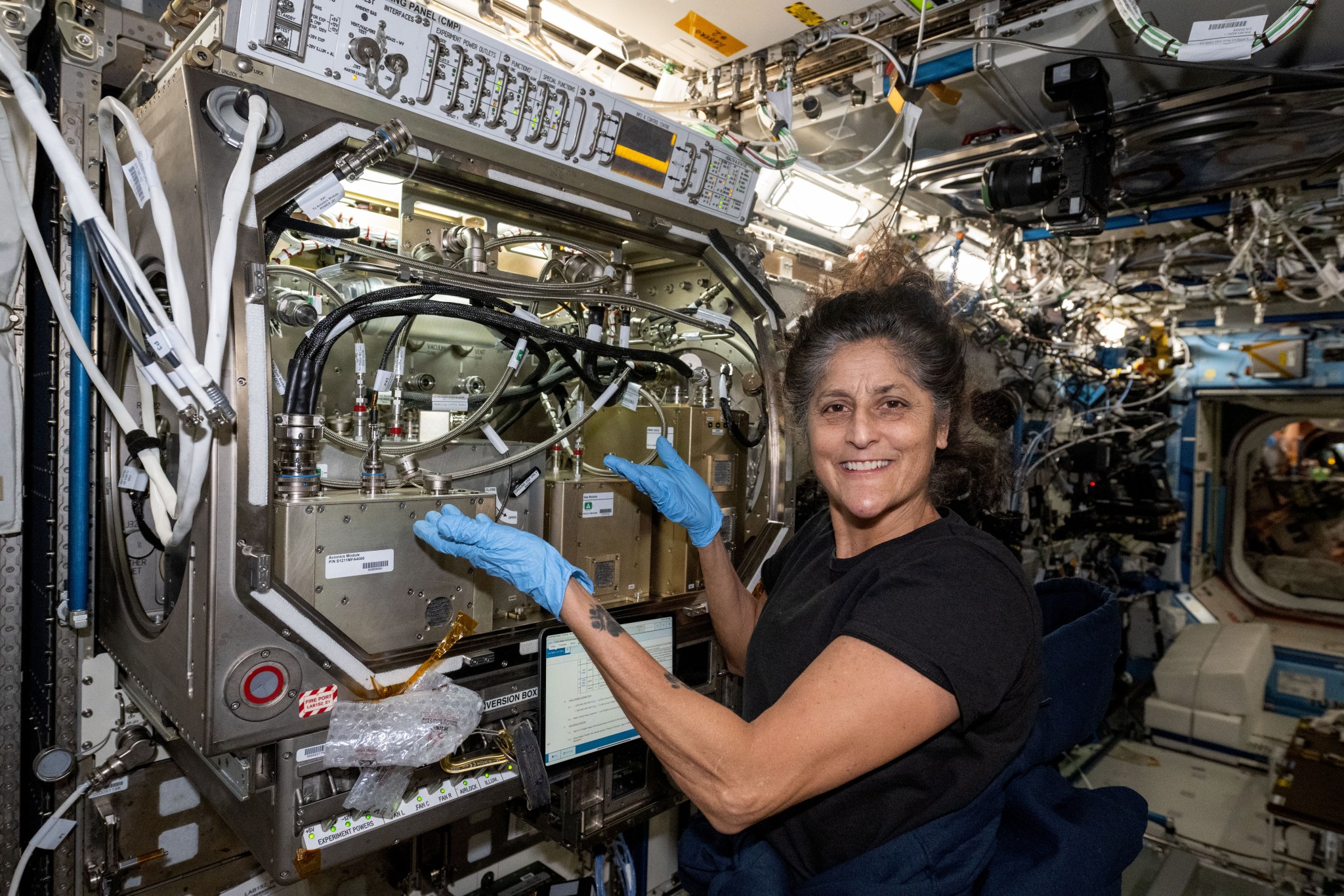 Williams, wearing a short-sleeved black t-shirt and blue latex gloves, smiles at the camera and gestures toward the Packed Bed Reactor in front of her. The front of the reactor has a silver box to her left and two gold boxes to her right, with multiple hoses and cords connecting them. There is a control panel across the top of the equipment with a row of switches and lights.