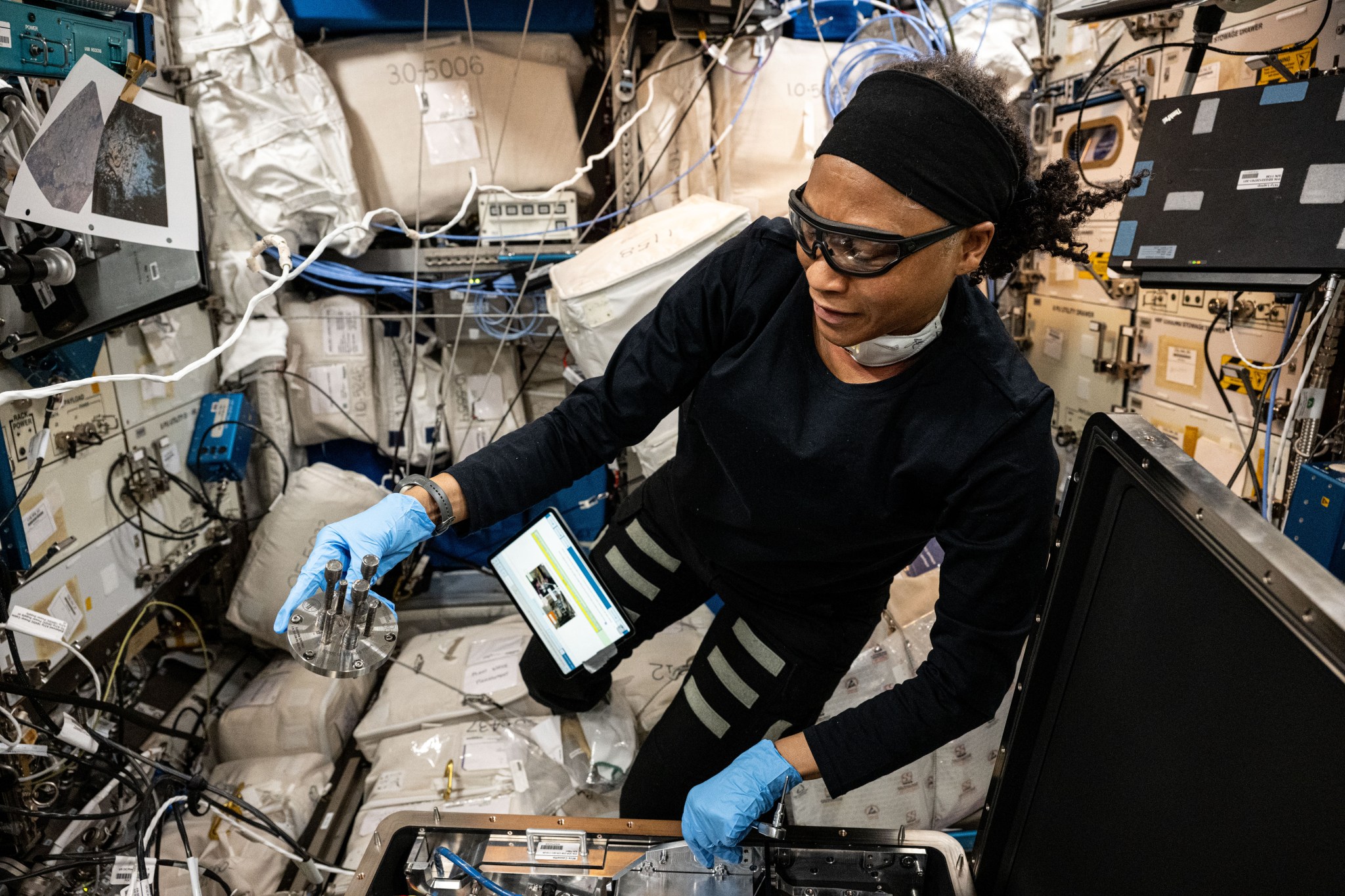 Epps is wearing a long-sleeved black top and pants, a black headband, goggles, and blue latex gloves and has a tablet attached by Velcro to her pants. In her right hand she holds a small metal disk with six 3D printed posts of different shapes and lengths protruding from it.