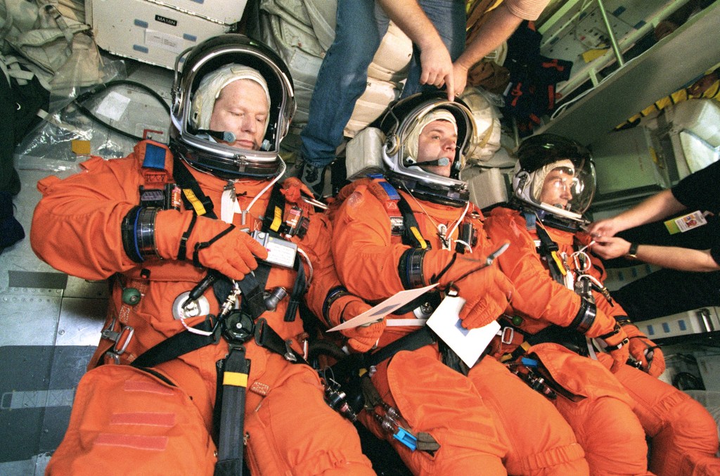 Three astronauts wear bright orange spacesuits, lying side by side inside a spacecraft or training module. The astronauts are suited up with their helmets on.